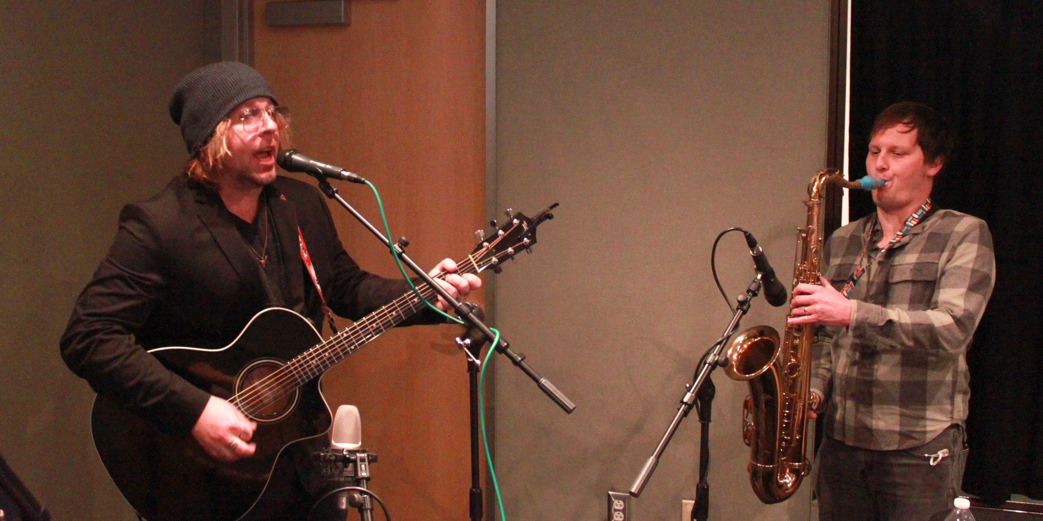 An adult playing guitar singing into a microphone and an adult playing tenor saxophone in a radio studio