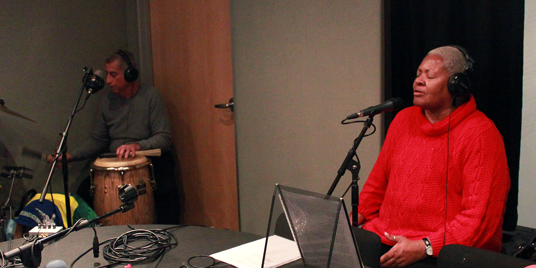 Two adults wearing headphones in a radio studio, one playing congas and cymbals, the other singing into a microphone