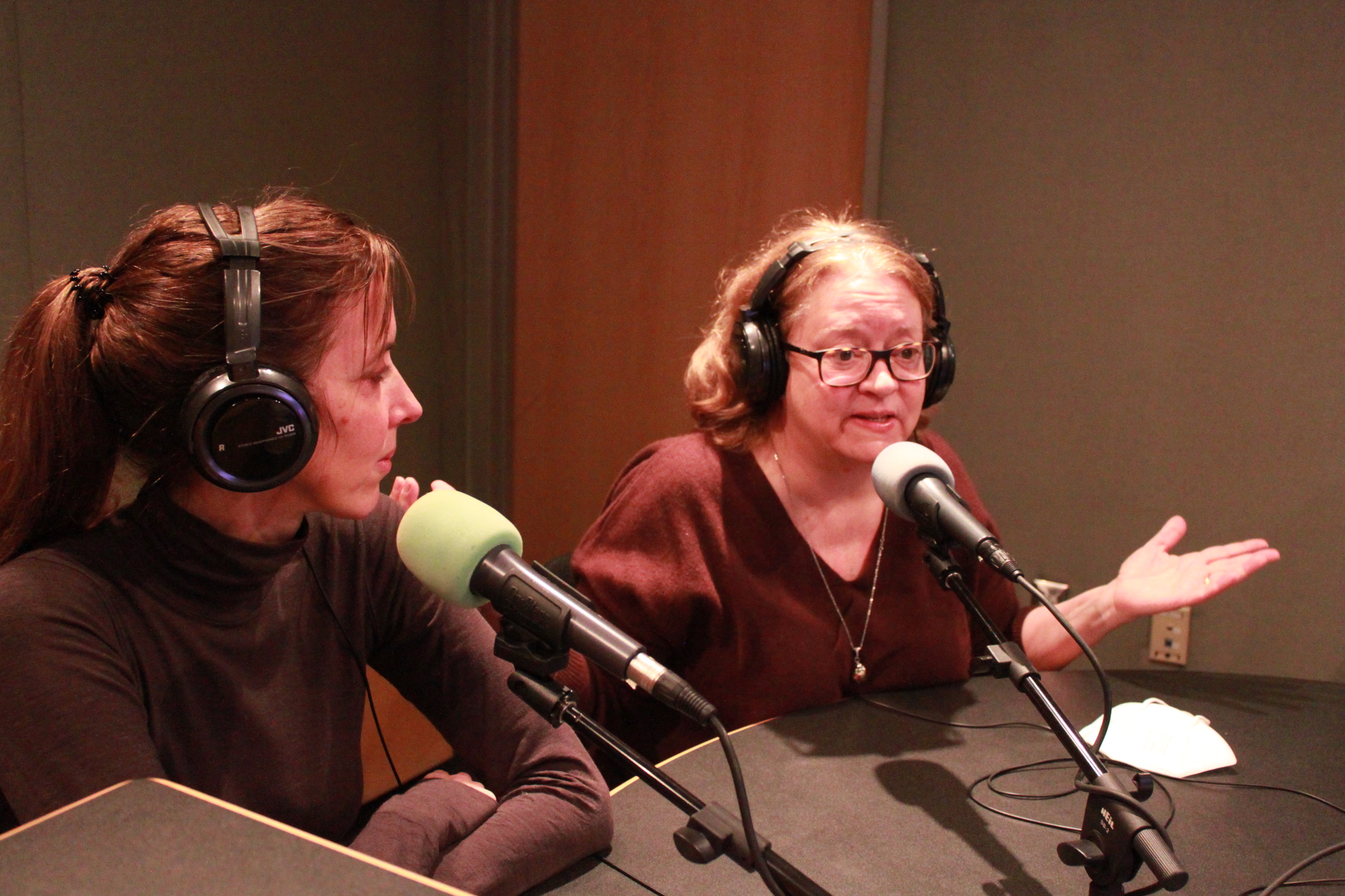 Two adults wearing headphones speaking into microphones in a radio studio