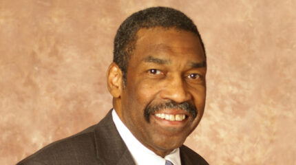 Adult with short graying hair, a short graying mustache, and a gray suit smiling in profile