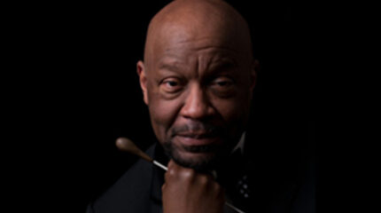 Bald adult with a short black mustache and beard dressed in a black suit and holding a conducting baton posing in profile