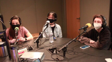 Three teens sitting in front of microphones, wearing headphones and face masks.