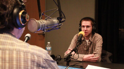 Adult wearing headphones speaking into a microphone being interviewed by an adult wearing headphones sitting behind a microphone in a radio studio