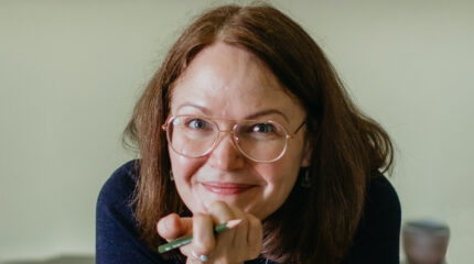 Adult with brown hair and glasses holding a pencil and smiling in profile