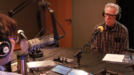 Person with gray hair and glasses wearing headphones and a plaid shirt talking into a microphone in a radio studio.