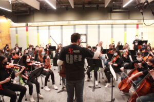 An adult wearing a black shirt conducting youth in an orchestra