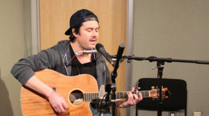 Adult wearing a navy backwards baseball cap playing guitar and singing into a microphone live on the radio