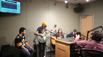 Four adult musicians in a radio studio performing live on two guitars, bass, and cajon .