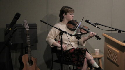 Person wearing glasses and a cream colored sweater playing a violin live on the radio.