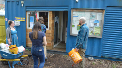 Four adults outside a blue building with buckets and a wheelbarrow