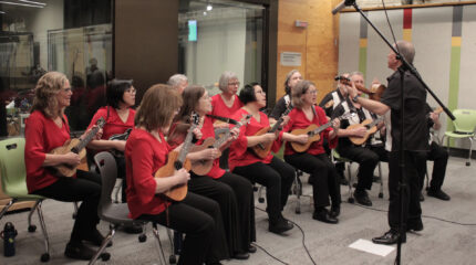 twelve adults playing ukuleles and one adult conducting while playing ukulele in an open room