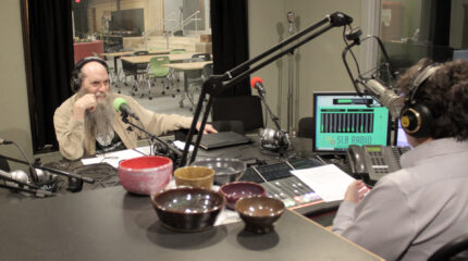 Bald bearded adult with headphones being interviewed in a radio studio, with several ceramic hand made bowls sitting nearby