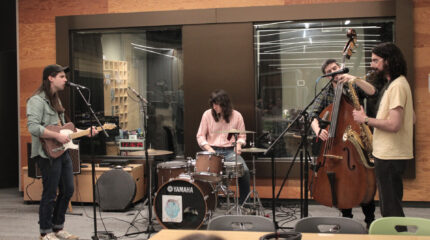 Four adults performing on guitar, drums, upright bass, and saxophone