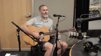 Adult playing guitar and singing into a microphone in a radio studio