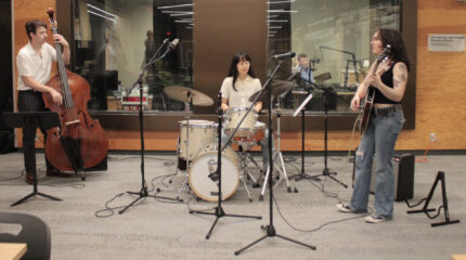 Three adult musicians performing in front of a radio studio on upright bass, drum set, and guitar