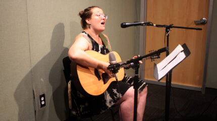 An adult playing guitar and singing into a microphone in front of sheet music on a music stand in a radio studio