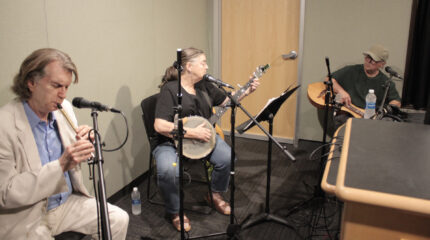 An adult playing tin whistle, an adult playing banjo, and an adult playing dulcimer in a radio studio