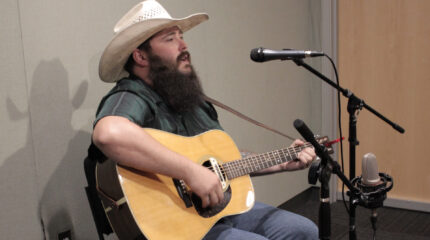 Adult with a tan wide brimmed hat playing guitar and singing into a microphone in a radio studio