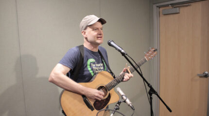 Adult playing guitar and singing into a microphone in a radio studio