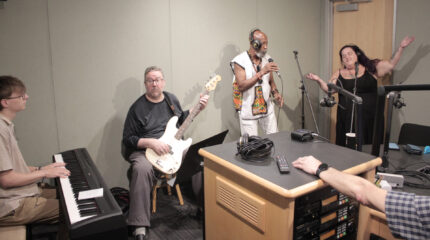 An adult playing piano keyboard, an adult playing bass, and two adults wearing headphones and singing into microphones in a radio studio