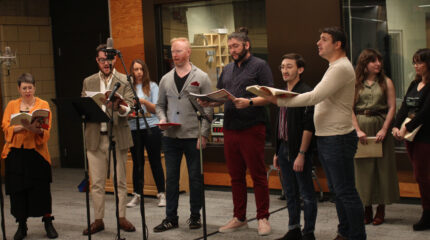 Nine adults holding scripts and singing into microphones