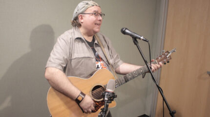 Adult playing guitar and singing into a microphone in a radio studio