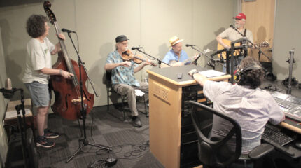 An adult playing upright bass, an adult sitting and playing fiddle, an adult sitting and playing banjo, and an adult playing acoustic guitar while another adult sitting and wearing headphones listens in a radio studio