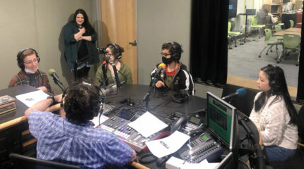 Four teens wearing headphones being interviewed by an adult wearing headphones siting behind a microphone and a mixing board while another adult stands in the corner listening in a radio studio