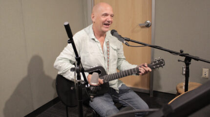 An adult singing into a microphone and playing guitar in a radio studio