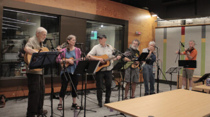 Two adults playing guitar, one adult playing ukulele, one adult playing mandolin, one adult playing harmonica, and an adult playing fiddle, all standing behind music stands