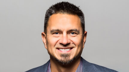 Adult with dark hair, a graying goatee, and a blue suit smiling in profile