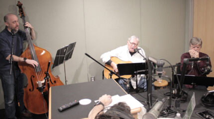 An adult playing upright bass, an adult playing guitar and singing into a microphone, and an adult playing harmonica into a microphone in a radio studio