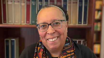 Adult with short graying hair, glasses, a multicolored scarf and a black top smiling in profile