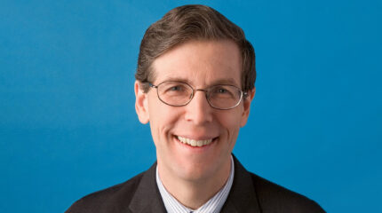 Adult with short brown hair, glasses, and a black suit smiling in profile