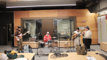 An adult playing bass, an adult playing a drum set, an adult paying guitar, and an adult playing ukulele and singing into a microphone