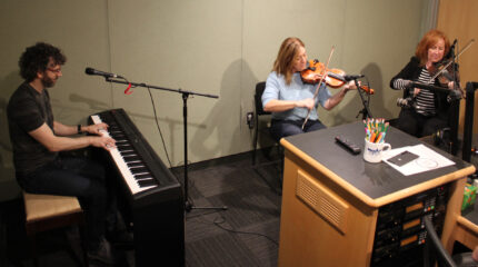 An adult playing a piano keyboard and two adults sitting and playing violins into microphones in a radio studio