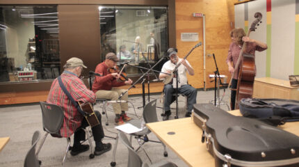 An adult sitting and playing guitar, an adult sitting and playing violin, an adult wearing headphones sitting and playing banjo, and an adult standing and playing upright bass, all playing into microphones