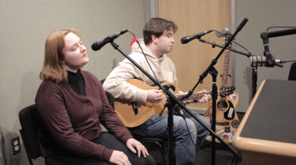 Two adults singing into microphones in a radio studio, one playing a guitar