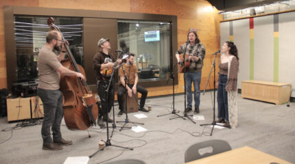 An adult playing upright bass, an adult playing mandolin and singing into a microphone, an adult playing cajon, an adult playing guitar and singing into a microphone, and an adult singing into a microphone