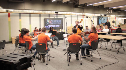 9 individuals playing string instruments in an orchestra while six adults watch