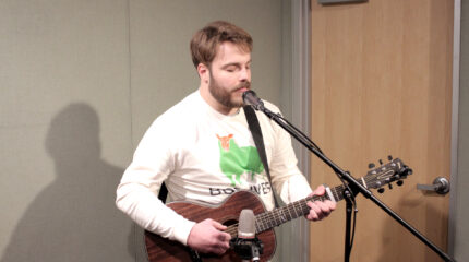 An adult playing an acoustic guitar and singing into a microphone in a radio studio