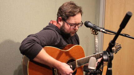An adult playing acoustic guitar sitting behind microphones in a radio studio