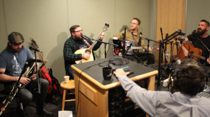 An adult playing an uilleann pipe, an adult playing bouzouki and singing into a microphone, an adult playing acoustic guitar, and an adult playing acoustic guitar and singing into a microphone, while an adult wearing headphones listens in a radio studio