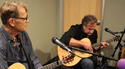 Two adults playing acoustic guitars sitting behind microphones in a radio studio