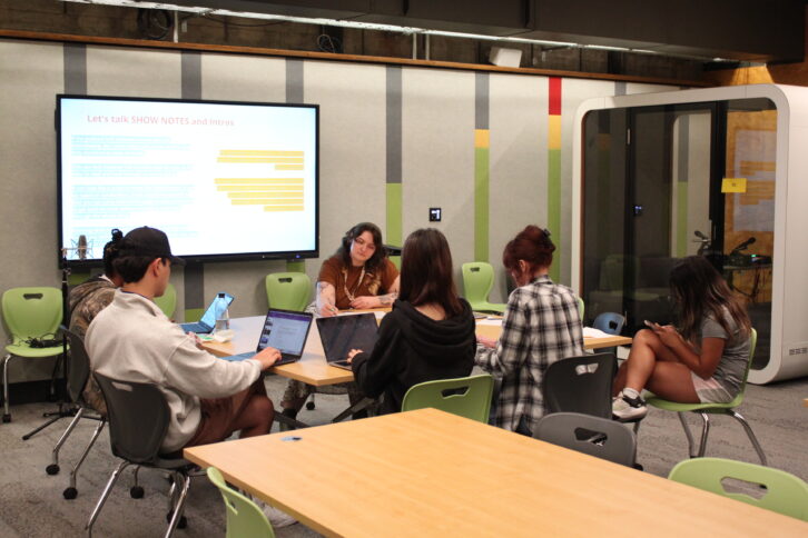 Four youth looking at laptops on a table, one youth looking at a smartphone, and one adult writing on a piece of paper on a table