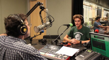 A teen wearing headphones speaking into a microphone being interviewed by an adult wearing headphones sitting behind a mixing board and a microphone in a radio studio
