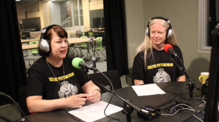 Two adults wearing headphones speaking into microphones in a radio studio
