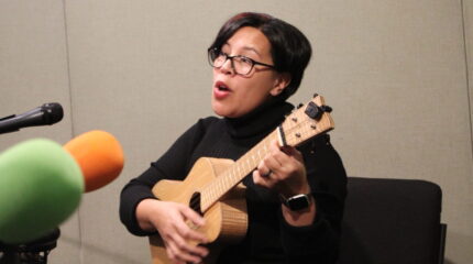 An adult playing ukulele and singing into a microphone in a radio studio
