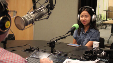 A teen wearing headphones sitting behind a microphone being interviewed by an adult wearing headphones sitting behind a microphone and a mixing board in a radio studio