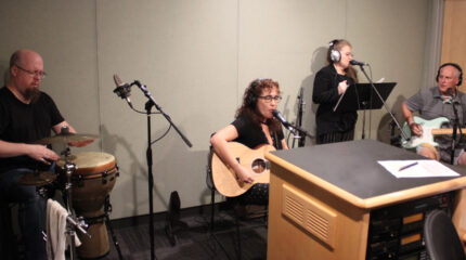 An adult playing percussion instruments, an adult playing acoustic guitar and singing into a microphone, an adult wearing headphones singing into a microphone, and an adult wearing headphones playing electric guitar in a radio studio